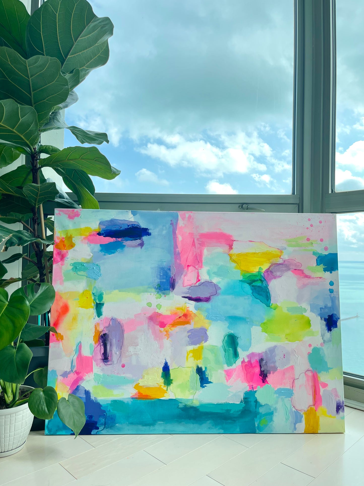 The Dolly Original Abstract Painting by Sarah Henry as part of the Happy Art Collection. The painting is shown on the light wooden floor of a studio leaning against a floor length window showing the blue clouds while several green plants sit beside it.