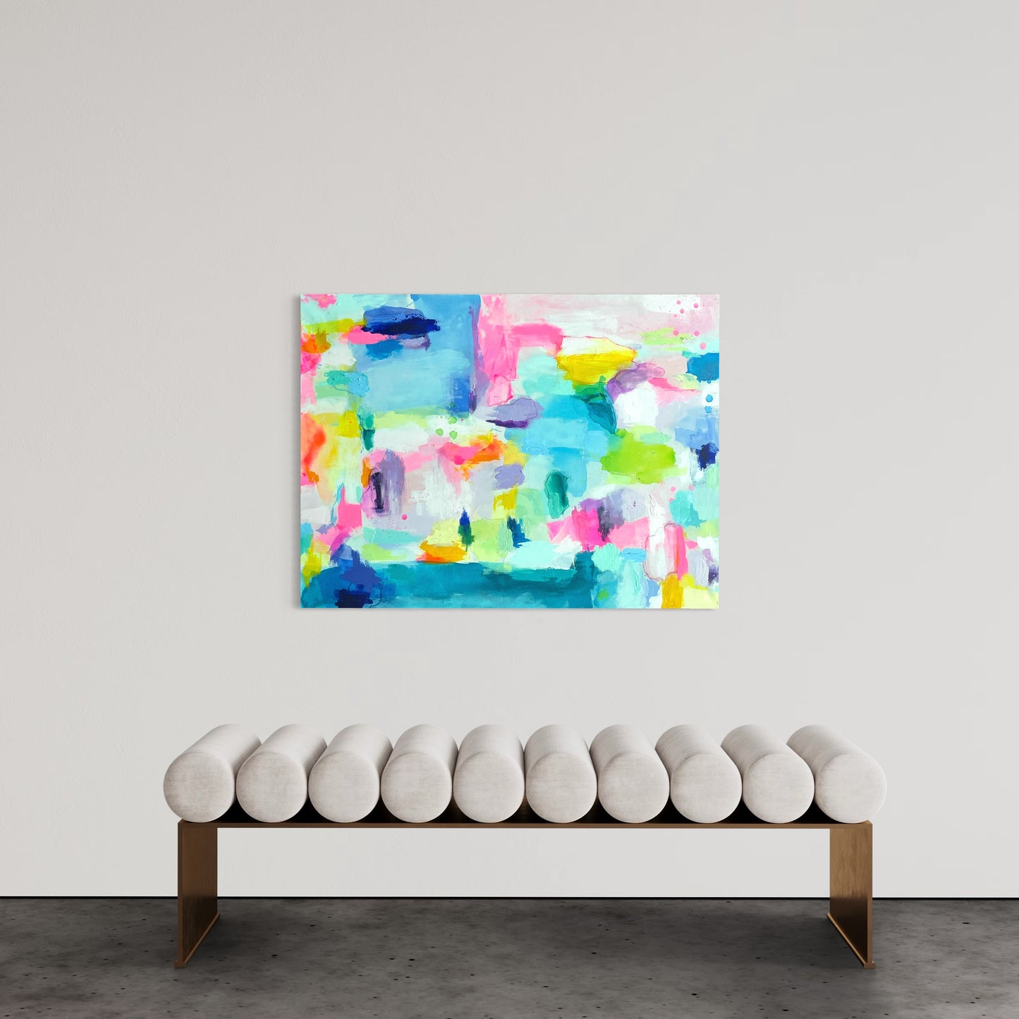 A contemporary and grand millennial interior features a large preppy abstract painting with vibrant colors, including shades of blue, pink, yellow, and green, hanging on a neutral-toned wall. The painting has an expressive, layered texture with bold brushstrokes. Below it, a modern bench with a sleek gold frame and cylindrical white cushions is positioned against the wall. The setting is minimalist, allowing the artwork to be the focal point of the space.