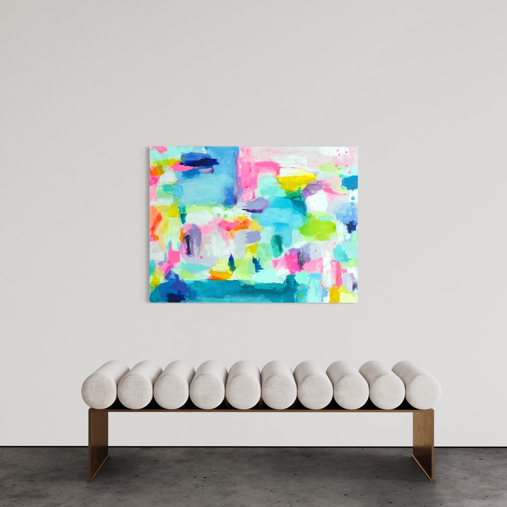 A contemporary and grand millennial interior features a large preppy abstract painting with vibrant colors, including shades of blue, pink, yellow, and green, hanging on a neutral-toned wall. The painting has an expressive, layered texture with bold brushstrokes. Below it, a modern bench with a sleek gold frame and cylindrical white cushions is positioned against the wall. The setting is minimalist, allowing the artwork to be the focal point of the space.