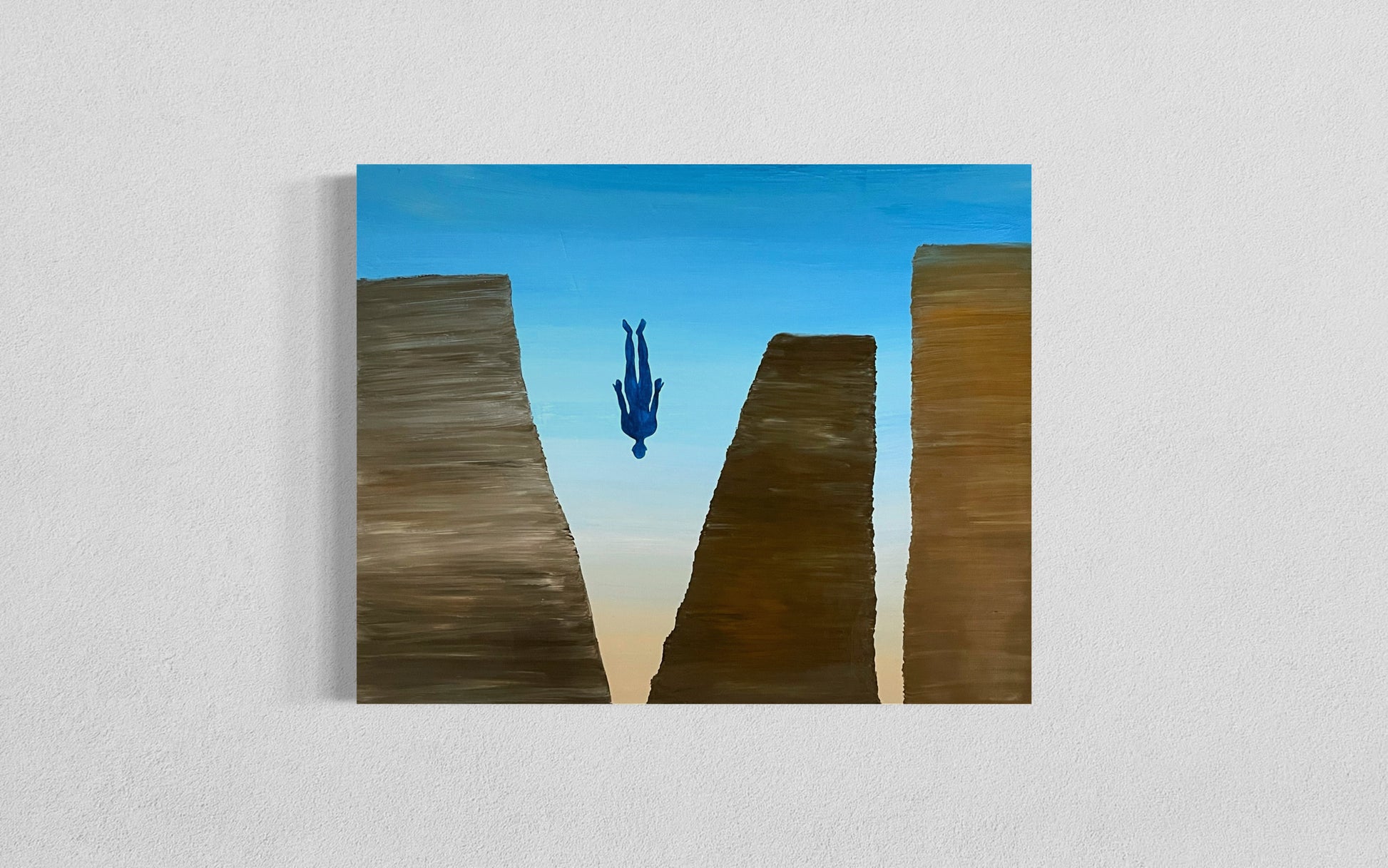 This surreal painting features a lone blue human figure falling headfirst between three towering rock formations. The background consists of a gradient sky transitioning from deep blue at the top to a warm golden hue near the horizon. The rock formations are textured in earthy browns, with the central one appearing smaller and more distant. The painting evokes a sense of isolation, uncertainty, or transcendence.