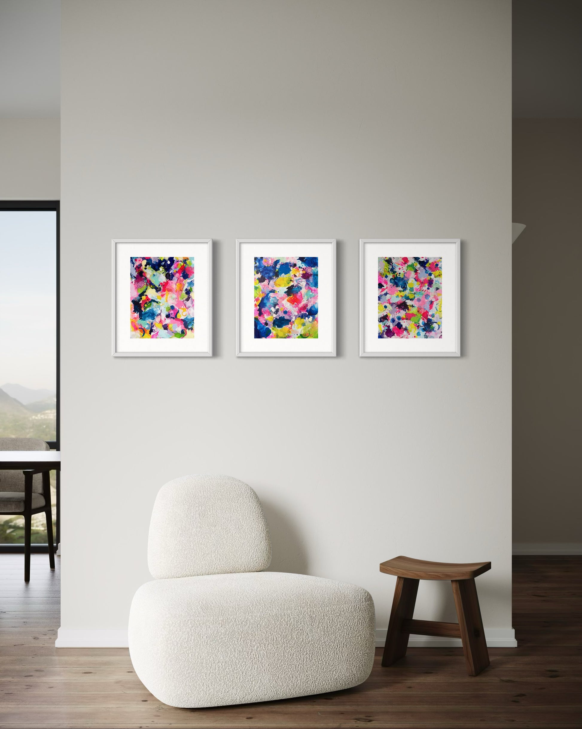 A modern collection of affordable abstract paintings featuring vibrant splashes of color, including pink, blue, yellow, green, purple and white. Each canvas is filled with expressive, energetic brushstrokes and layered paint splatters, creating a dynamic and contemporary composition. The modern wall decor is arranged side by side on a white wall in a hallway.