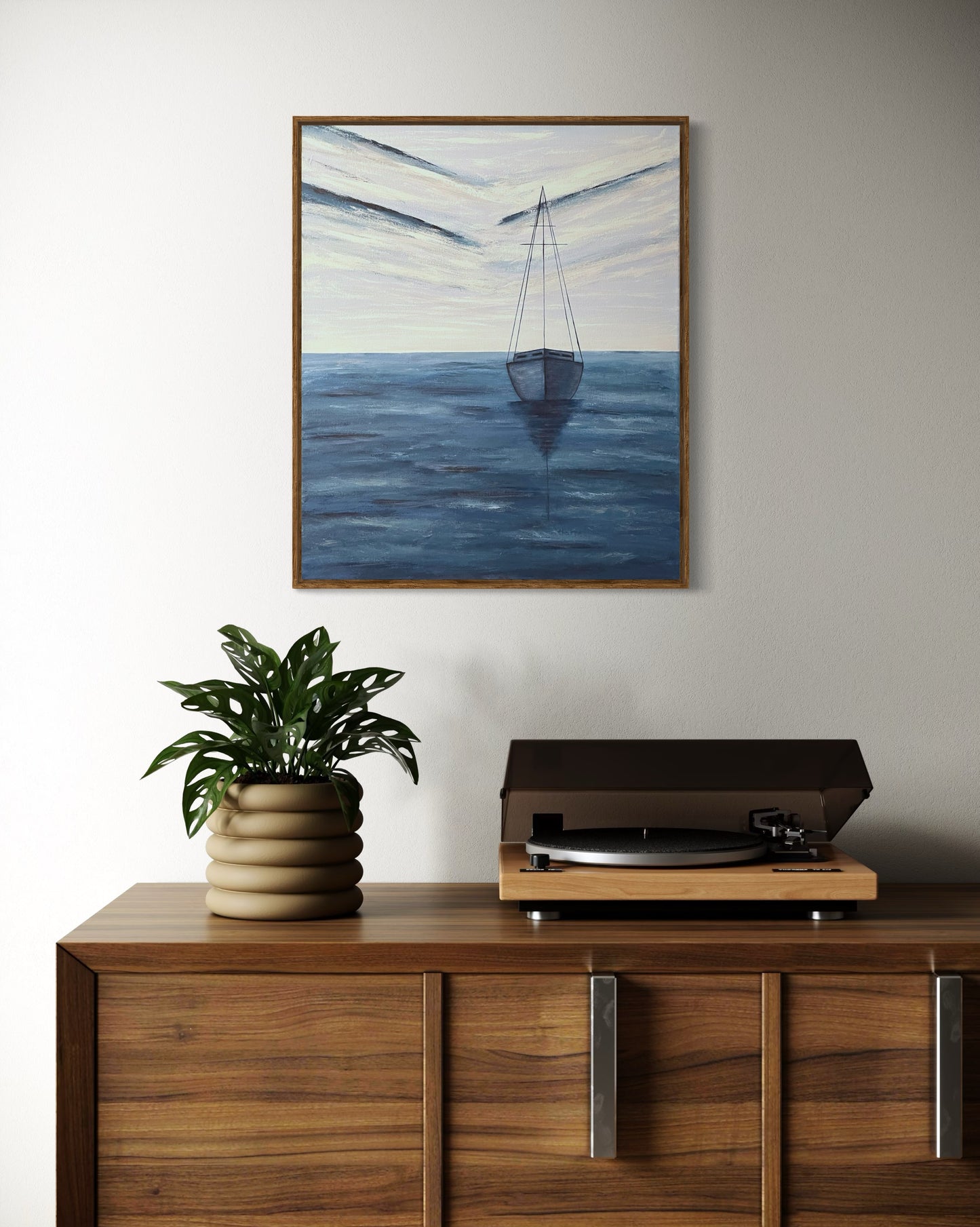 An affordable serene ocean painting of a solitary sailboat floating on calm, deep blue waters. The boat is slightly off center in the composition, with its reflection subtly visible on the water’s surface. The sky above is painted with soft, muted tones of pale blue, white and navy, with expressive brushstrokes forming abstract cloud patterns. The horizon line is faint, blending seamlessly with the sky and sea, creating a tranquil and minimalist atmosphere. The wall decor hangs on a white wall in a hallway.