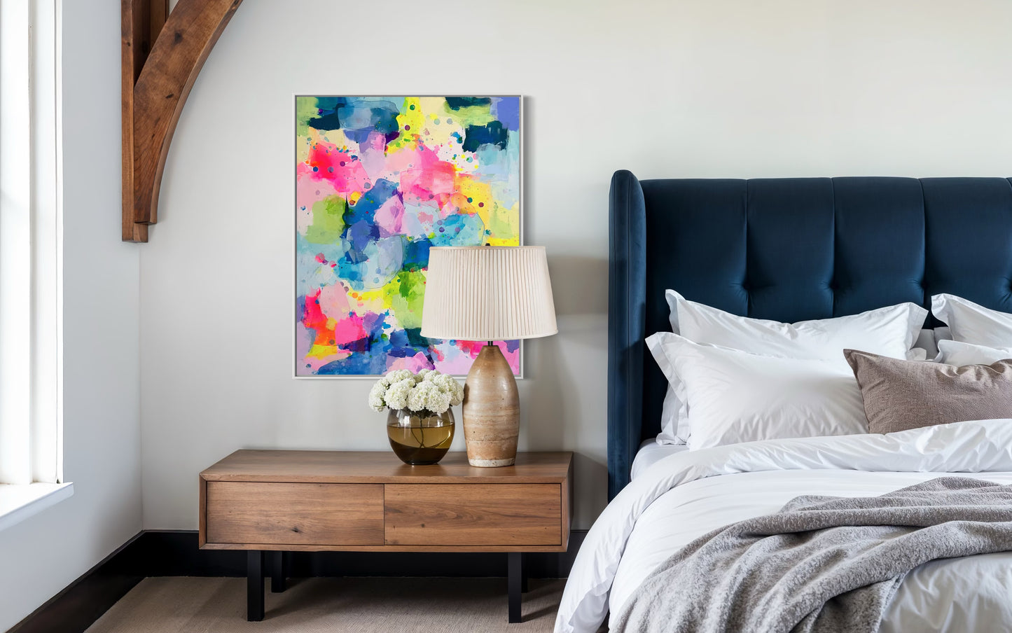 A happy, preppy affordable abstract painting featuring vibrant splashes of pink, blue, yellow, and green. The fluid brushstrokes and playful color combinations create a cheerful, energetic art composition with a watercolor-like effect. The layering of translucent hues and scattered paint splatters evoke a sense of lightness and spontaneity, embodying a bright and uplifting aesthetic for a dopamine boost. This preppy colorful maximalism wall decor hangs on a white bedroom wall next to a navy velvet bed.