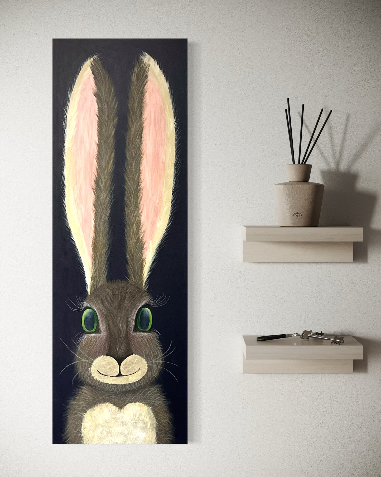 A 12"x36" oblong abstract acrylic painting with a cartoon style hare rabbit bunny with dazzling green eyes, gray and brown fur, pink ears, long eyelashes, on a navy blue background hangs on a white wall next to minimalistic style shelves.