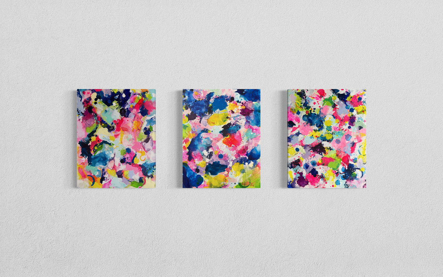 A modern gallery collection of abstract paintings featuring vibrant splashes of color, including pink, blue, yellow, green, purple and white. Each canvas is filled with expressive, energetic brushstrokes and layered paint splatters, creating a dynamic and contemporary composition. The affordable wall decor is arranged side by side on a white wall, forming a cohesive yet varied visual experience.