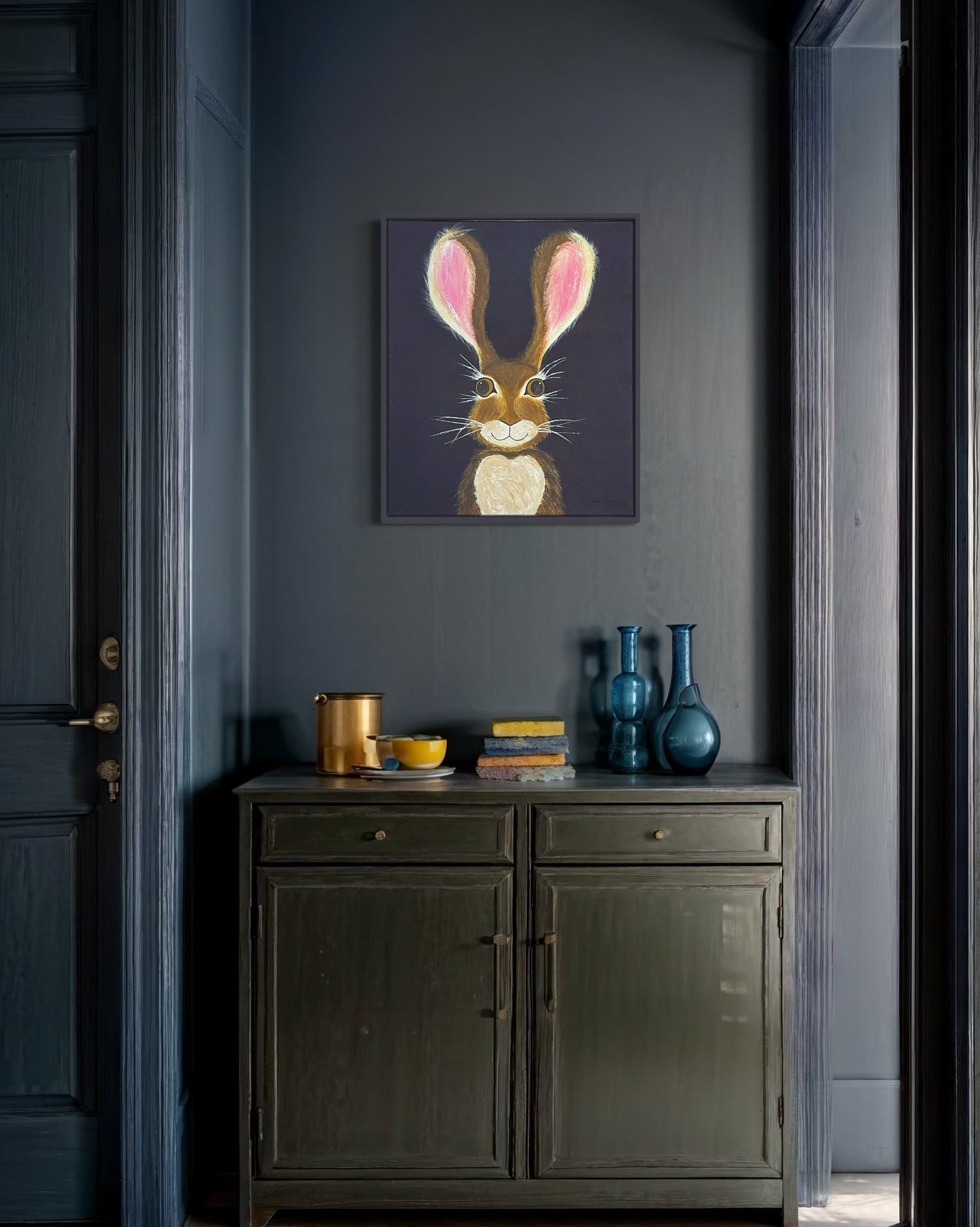 A 20"x24" affordable abstract acrylic painting with a cartoon style hare rabbit bunny with dazzling gold eyes, brown fur, pink ears, long eyelashes, on a navy blue background hangs on a blue wall in a hallway, entryway, office, or bedroom.