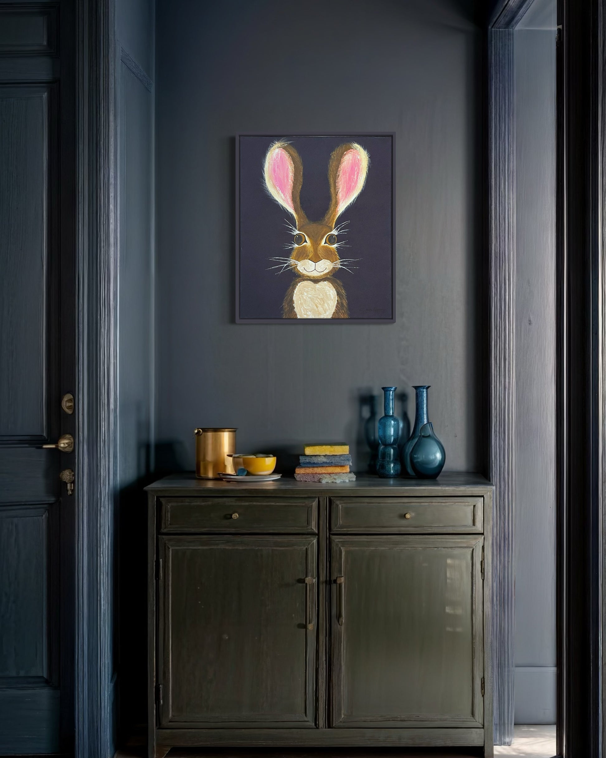 A 20"x24" affordable abstract acrylic painting with a cartoon style hare rabbit bunny with dazzling gold eyes, brown fur, pink ears, long eyelashes, on a navy blue background hangs on a blue wall in a hallway, entryway, office, or bedroom.