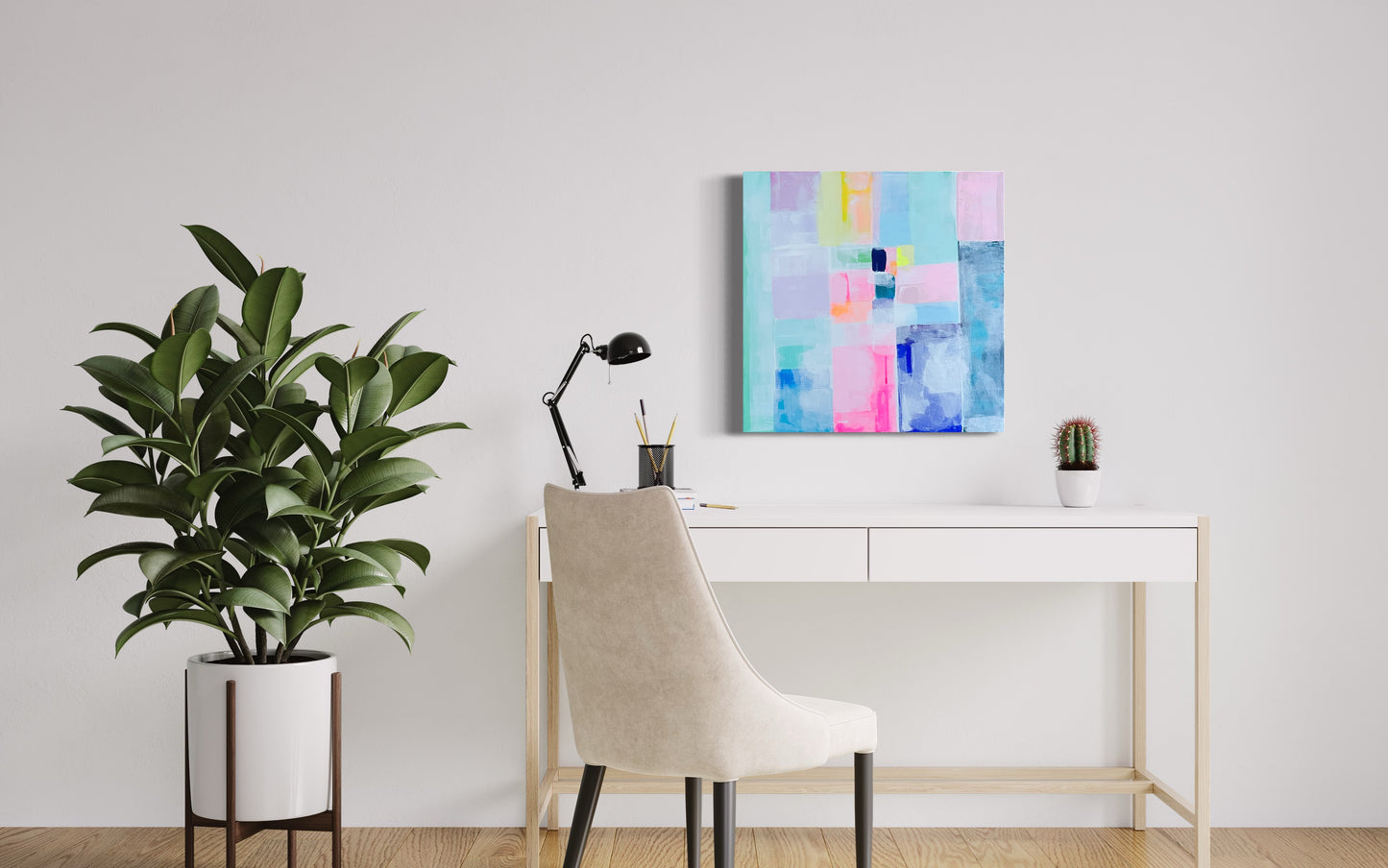 Modern minimalist home office with a pastel abstract geometric painting titled "A New Day" hanging above a sleek white desk; decor includes a beige chair, black lamp, cactus plant, and tall rubber plant in a white pot on wood flooring.