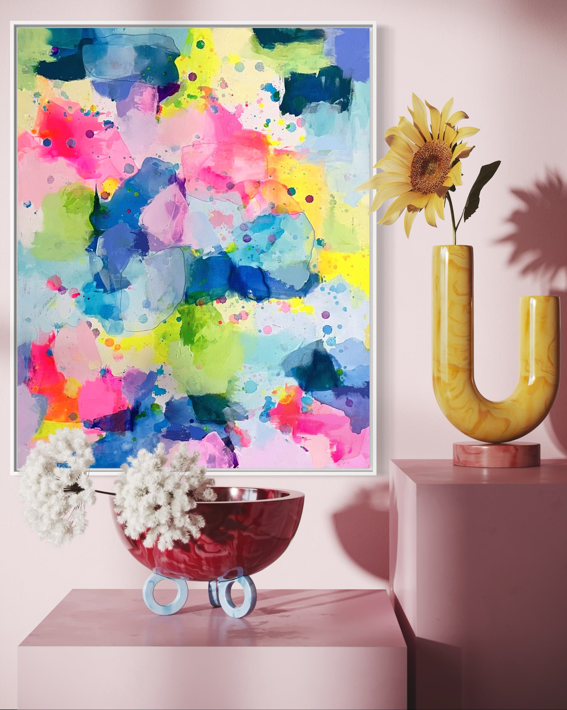 A happy, preppy affordable abstract painting featuring vibrant splashes of pink, blue, yellow, and green. The fluid brushstrokes and playful color combinations create a cheerful, energetic art composition with a watercolor-like effect. The layering of translucent hues and scattered paint splatters evoke a sense of lightness and spontaneity, embodying a bright and uplifting aesthetic for a dopamine boost. This preppy colorful maximalism wall decor hangs on a pink wall in an entryway or hallway.