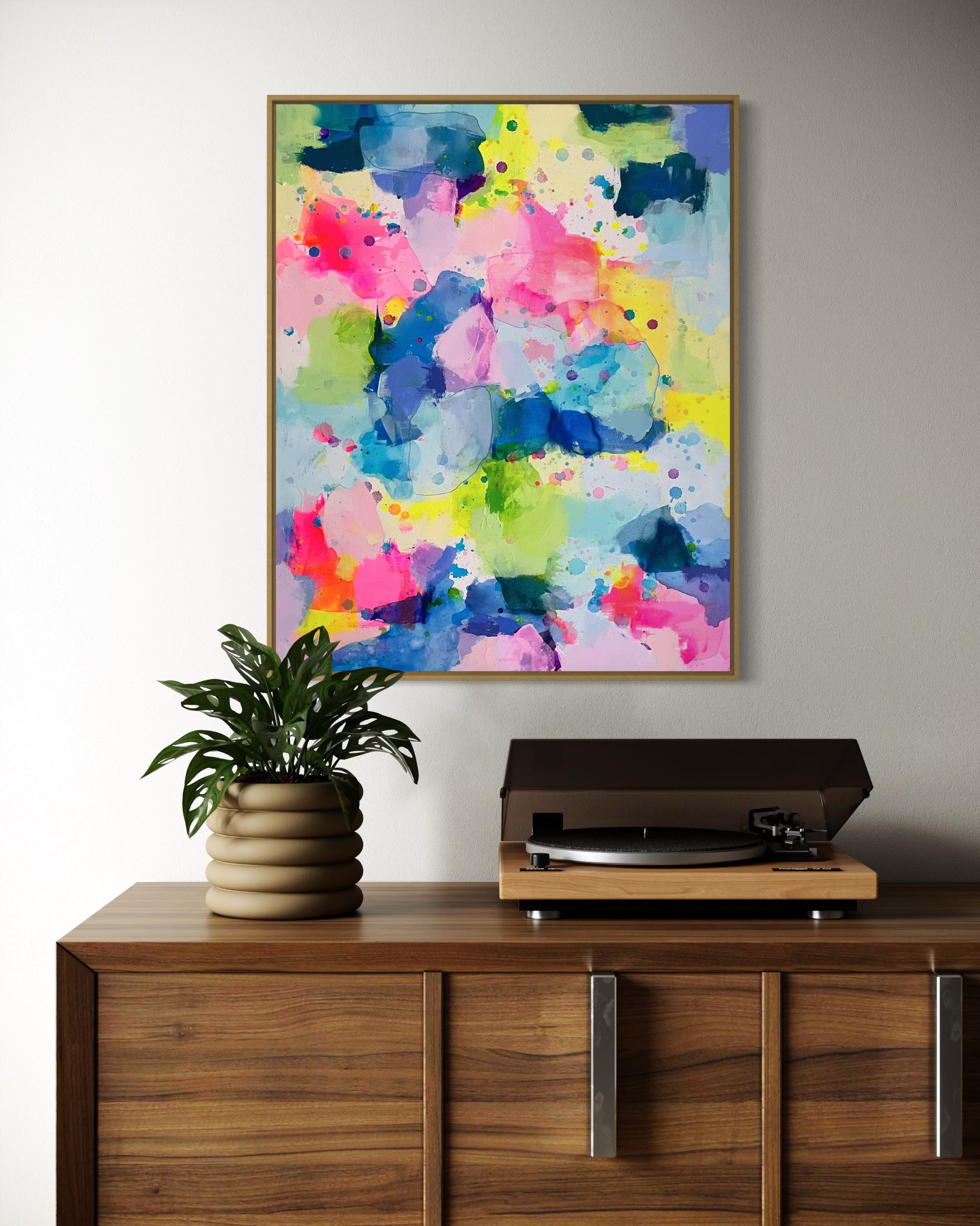 A happy, preppy affordable abstract painting featuring vibrant splashes of pink, blue, yellow, and green. The fluid brushstrokes and playful color combinations create a cheerful, energetic art composition with a watercolor-like effect. The layering of translucent hues and scattered paint splatters evoke a sense of lightness and spontaneity, embodying a bright and uplifting aesthetic for a dopamine boost. This preppy colorful maximalism wall decor hangs on a white wall in a living room.
